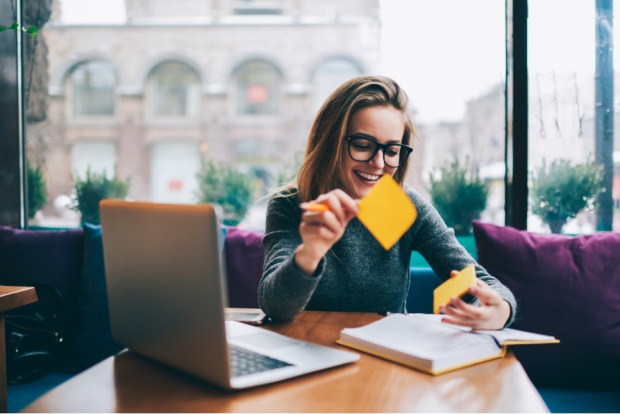 Junge Studentin mit Notebook, aufgeschlagenem Lehrbuch und Infokärtchen in einem städtischen Café | Credit: iStock.com/GaudiLab