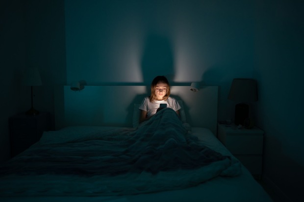 Eine Frau, die in einem dunklen Schlafzimmer in ihrem Bett sitzt und auf ihr Handy, dessen Bildschirmlicht ihr Gesicht erhellt, starrt