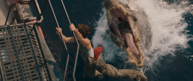 Szene aus "Jurassich World - Die Wiedergeburt", in der ein Dinosaurier mit weit aufgerissenem Maul aus dem Wasser springt und nach einem Mann schnappt, der sich an dem Geländer einer Brücke festklammert