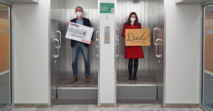 Ein Mann steht mit einem Spendenscheck in der Hand in einem Paternoster neben einer Frau, die ein Schild mit der Aufschrift "Danke" in Händen hält