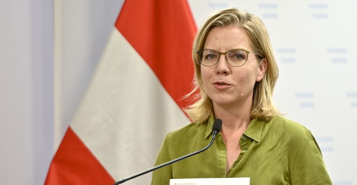 Leonore Gewessler bei einer Pressekonferenz. Sie steht vor einer Österreichfahne und spricht in ein Mikrofon.