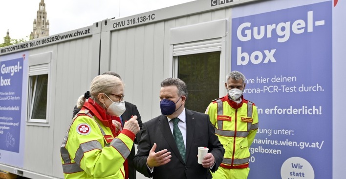 Bürgermeister Michael trägt eine Maske. Er steht mit zwei Sanitätern vor einer Gurgelbox.