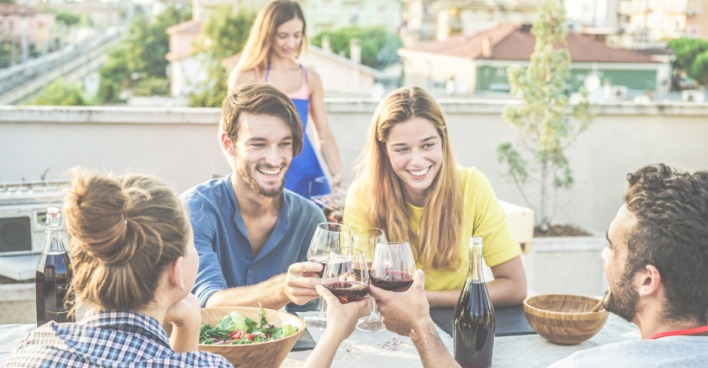 Junge Menschen genießen eine gemeinsame Mahlzeit unter freiem Himmel auf einer Dachterrasse | Credit: iStock.com/DisobeyArt