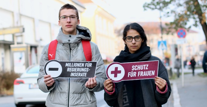 Ein junger Mann und eine junge Frau halten zwei Schilder mit Botschaften für mehr Gewaltschutz für Frauen in die Kamera