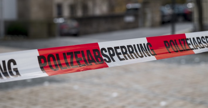 Ein rot-weiß-rotes Absperrband mit dem Schriftzug "Polizeiabsperrung" auf eine Straße mit Kopfsteinpflaster.