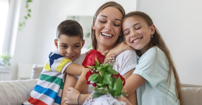 Sohn und Tochter umarmen ihre Mutter, die rote Rosen hält | Credit: istock.com/stefanamer