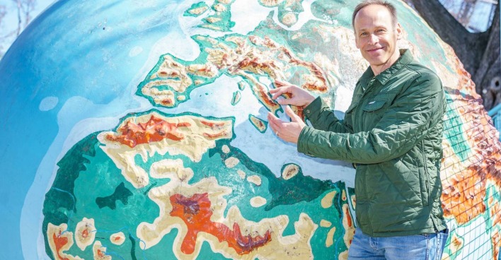 Meteorologe Marcus Wadsak steht vor einer Weltkugel und zeigt mit dem Finger auf ein Land