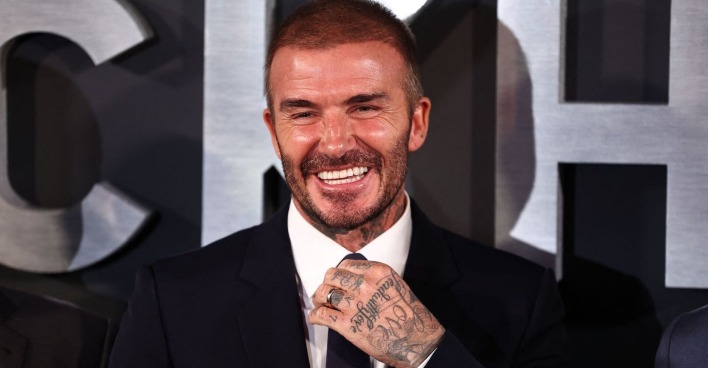 David Beckham lacht in die Kamera und richtet seine Krawatte.