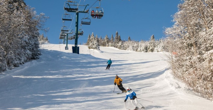 Drei Skifahrer auf einer Piste, blauer Himmel, darüber ein Sessellift.