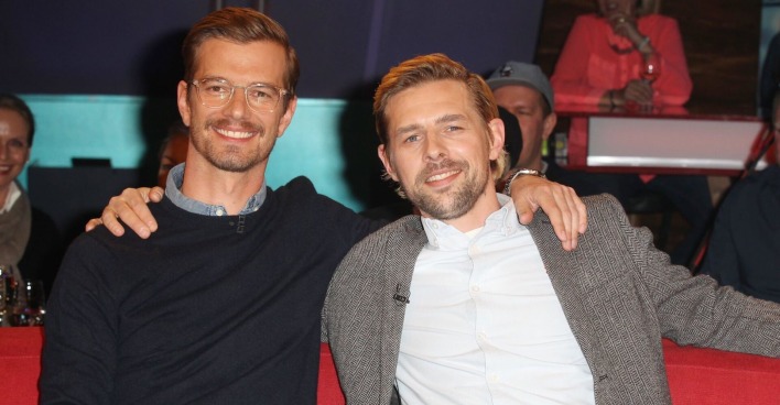 Joko Winterscheidt und Klaas Heufer Umlauf sitzen auf einer roten Bank und haben die Arme umeinander gelegt. Beide lachen in die Kamera.