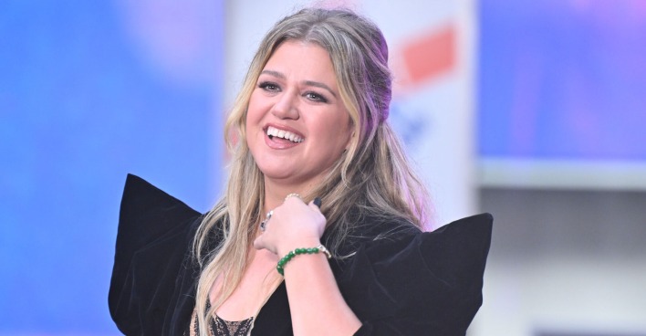 Kelly Clarkson trägt ein schwarzes Kleid und halb geschlossene blonde Haare. Sie lächelt in die Ferne.