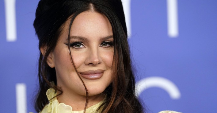 Lana Del Rey trägt hochgesteckte braune Haare und ein starkes Augen Make-Up. Sie steht vor einer blauen Wand.