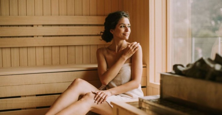 Eine Frau sitzt mit einem Handtuch bedeckt in einer gemütlichen Sauna aus Holz und schaut glücklich aus dem Fenster neben ihr.