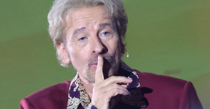 Thomas Gottschalk hält sich den Zeigefinger schweigend vor den Mund. Er trägt ein weinrotes Sakko.