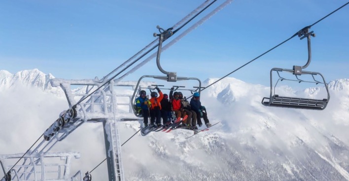 Ein Sessellift mit acht Skifahrern in der Luft, im Hintergrund eine schneebedeckte Gebirgskette