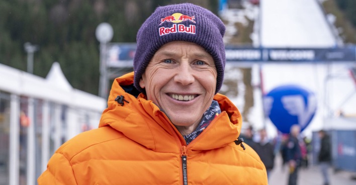 Ein Mann in oranger Skijacke und mit "Red Bull"-Haube steht vor einer Skipiste