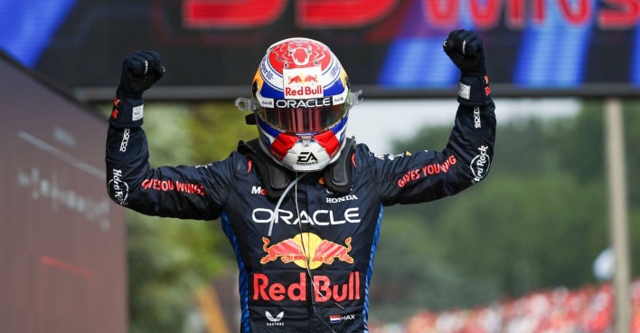 Max Verstappen, der im Zielbereich eines Grand Prix eine Bodybuilder-Geste vollführt und dabei seinen Rennhelm auf dem Kopf trägt