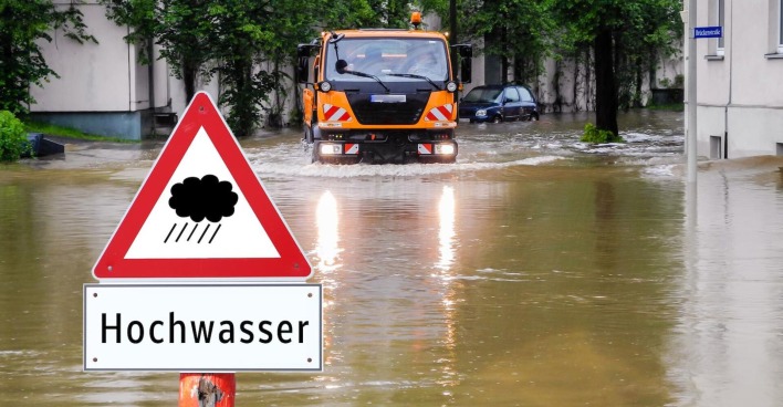 Hochwasser: Gemeinde-Lastwagen auf überschwemmter Straße und ein Hinweisschild "Hochwasser".
