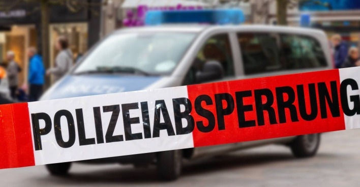 Ein Absperrband auf dem der Schriftzug "Polizeiabsperrung" zu lesen ist und in dessen Hintergrund sich ein leicht verschwommen erscheinendes Polizeiauto befindet