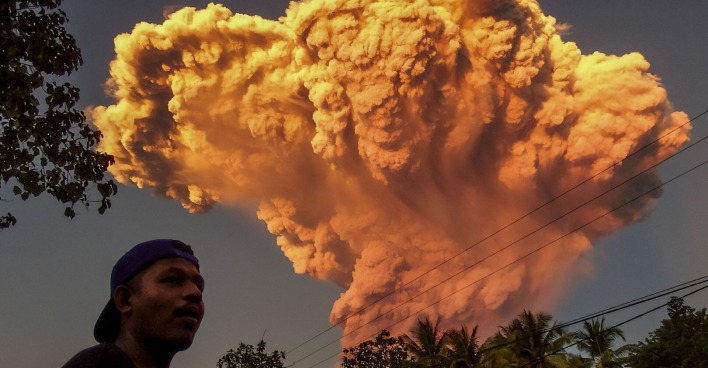 Ein Mann steht im Vordergrund, während im Hintergrund eine gewaltige orangefarbene Aschewolke vom ausbrechenden Vulkan Lewotobi Laki-Laki auf der indonesischen Insel Flores aufsteigt.