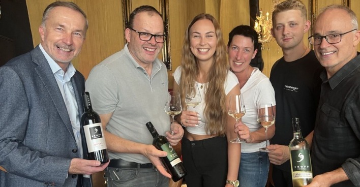 Vier Männer und zwei Männer lachen in die Kamera. In den Händen halten sie Weinflaschen beziehungsweise Weingläser, die mit Weißwein gefüllt sind.