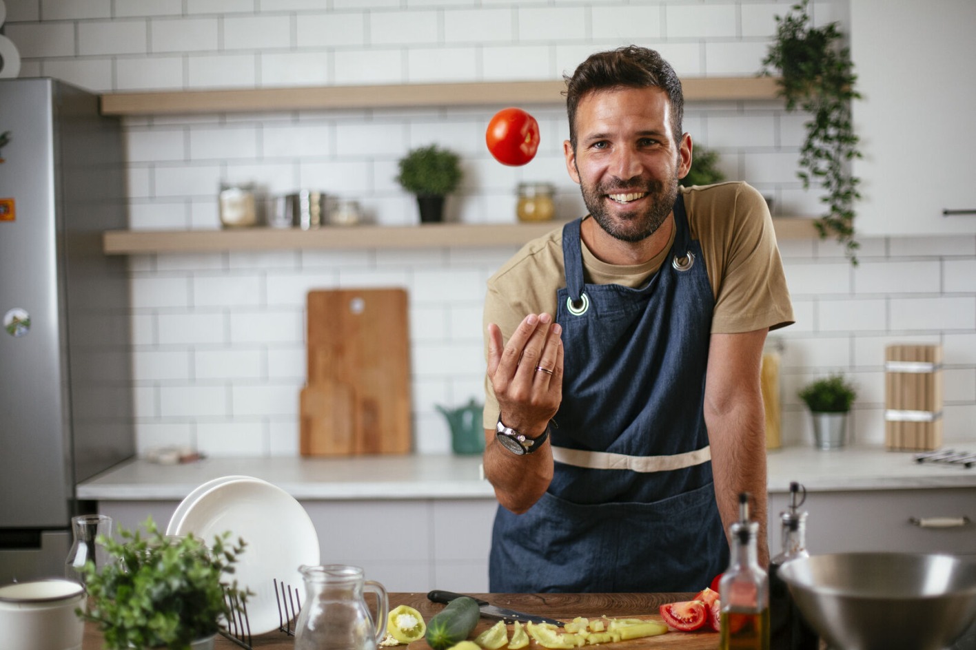 Ein Mann steht vor einem Küchentisch voller gesunder Lebensmittel und wirft lässig eine Tomate in die Höhe