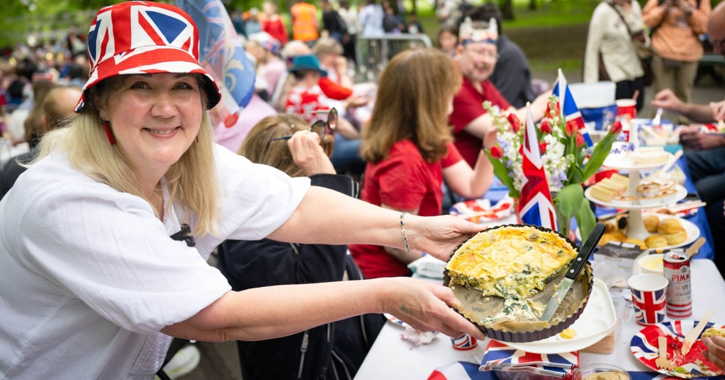 Eine Frau hält eine angeschnittene Coronation Quiche in die Kamera. Im Hintergrund viele Feiernde, sie trägt einen Hut mit der britischen Flagge.