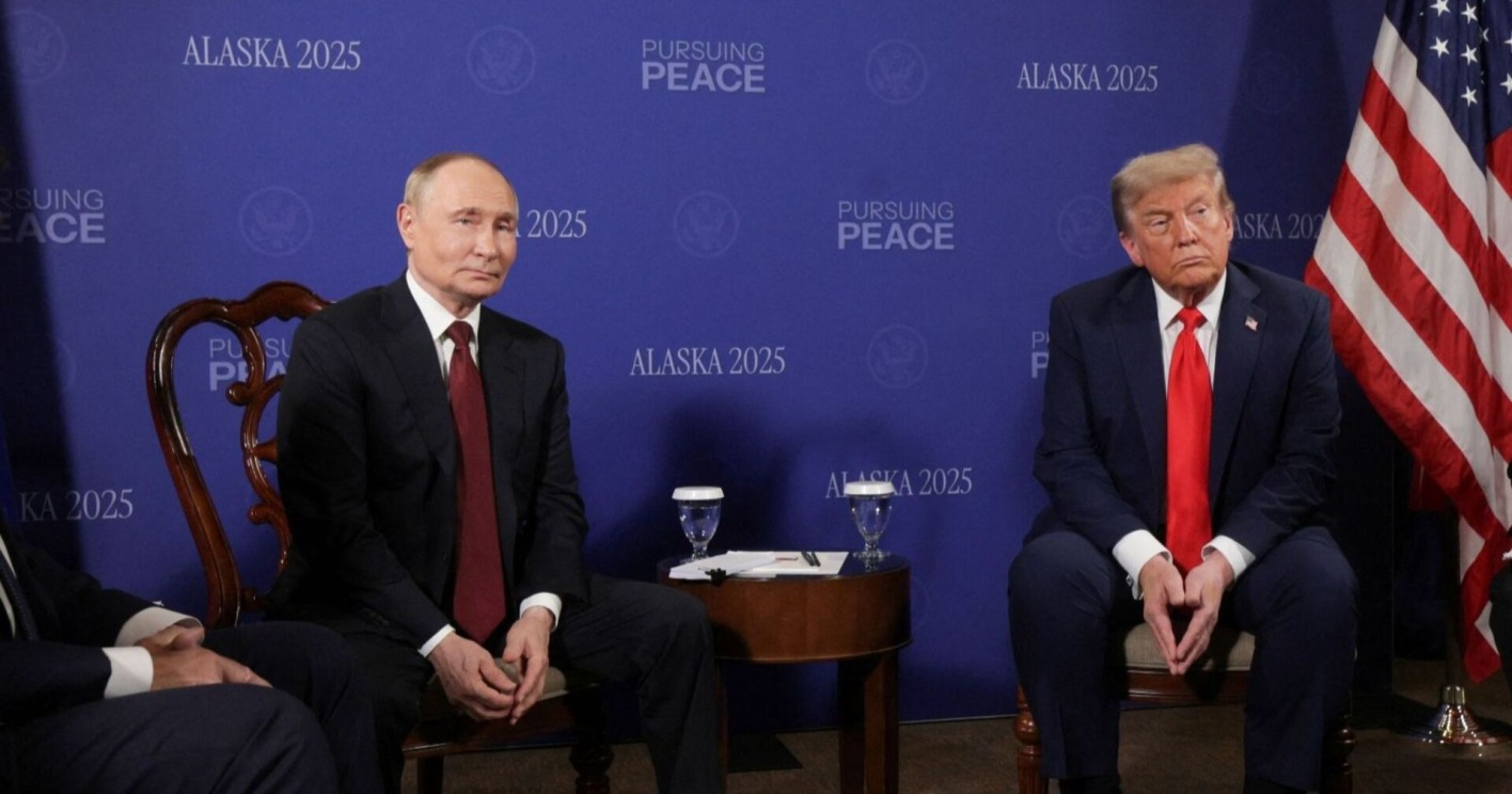 Donald Trump und Wladimir Putin sitzen nebeneinander vor blauem Hintergrund mit der Aufschrift „Alaska 2025“ und „Pursuing Peace“. Beide wirken ernst.