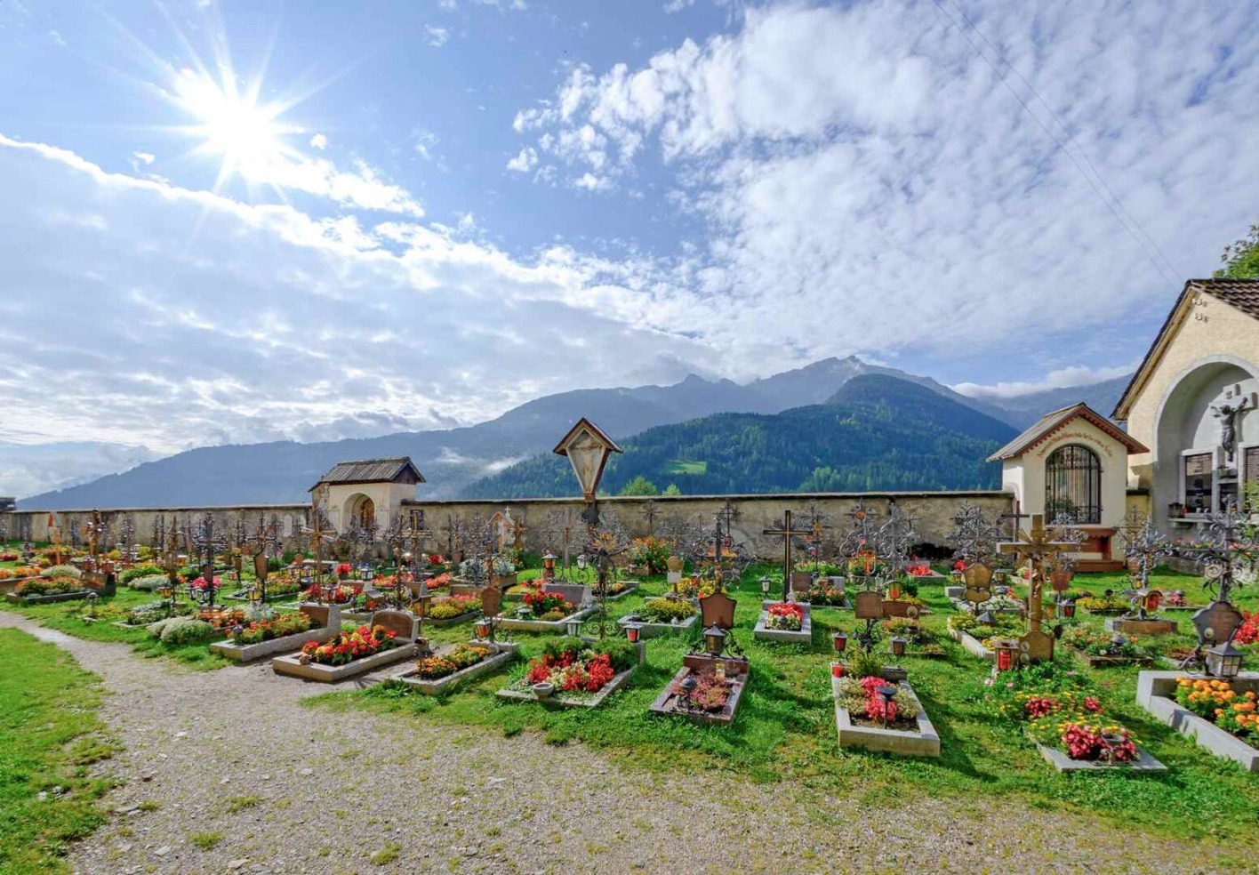 Blumenreicher Friedhof in den österreichischen Alpen bei Sonnenschein, mit Bergen im Hintergrund.