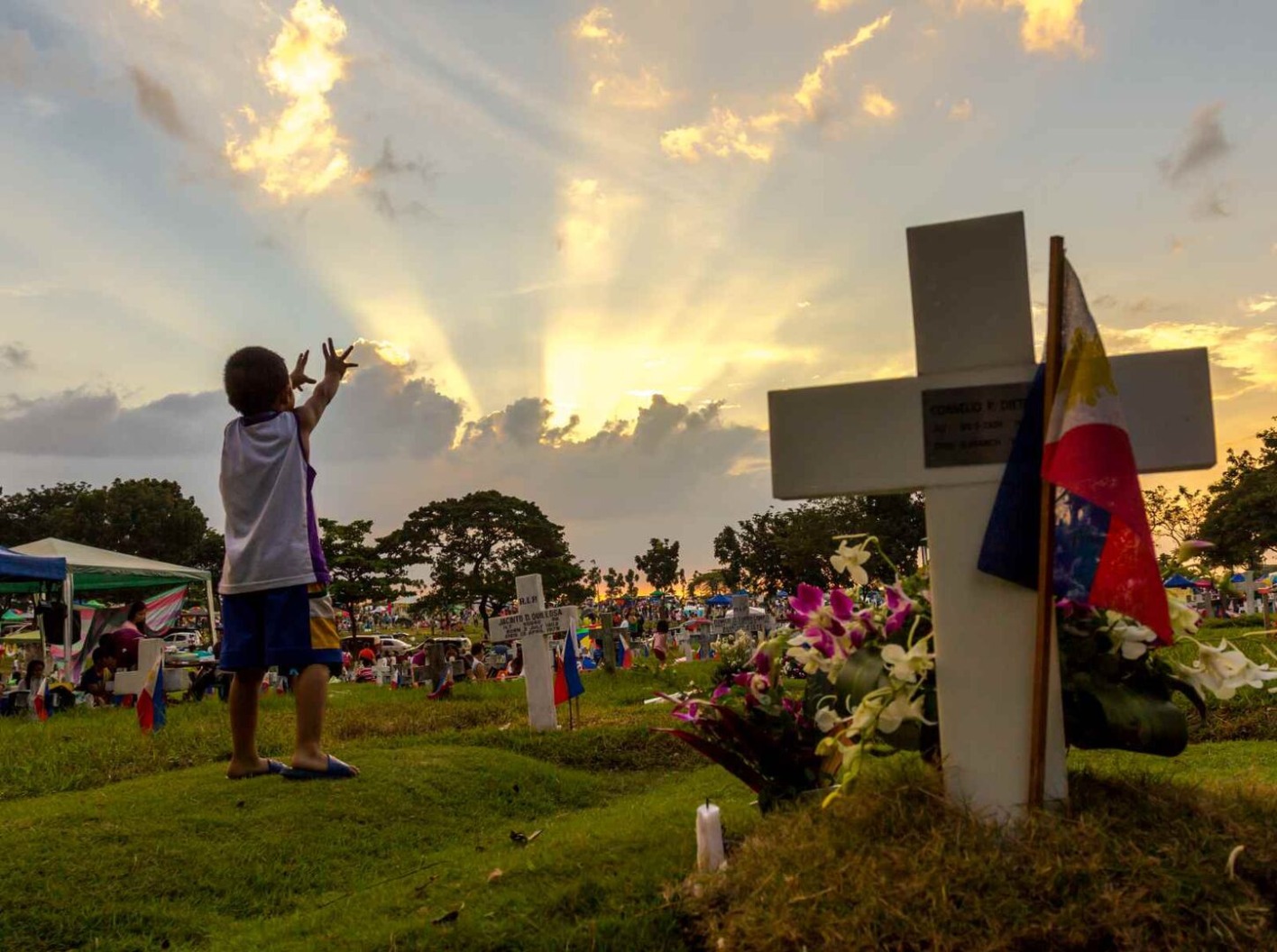 Kind auf einem Friedhof auf den Philippinen, das bei Sonnenuntergang an einem Grab mit Blumen und Flagge steht.