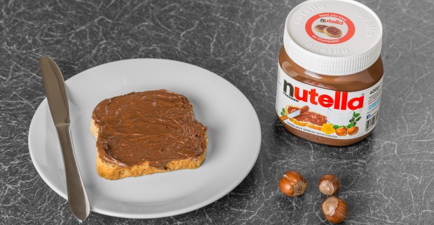 Mit Nutella beschmiertes Weißbrot neben einem Nutella-Glas und drei Haselnüssen