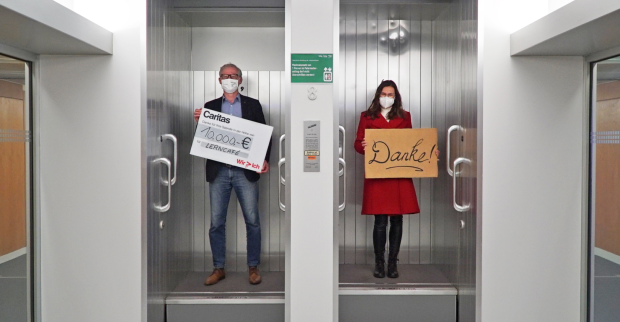 Ein Mann steht mit einem Spendenscheck in der Hand in einem Paternoster neben einer Frau, die ein Schild mit der Aufschrift "Danke" in Händen hält
