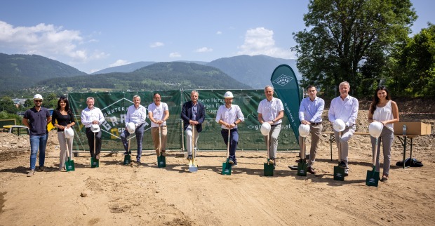 Eine Gruppe von Männern und Frauen steht auf Spaten gestützt auf einem Baugrundstück mit Panoramablick
