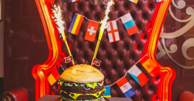 Eine Torte, die aussieht wie ein Burger mit sprühenden Wunderkerzen vor einem Ledersessel, der mit Flaggen geschmückt ist.