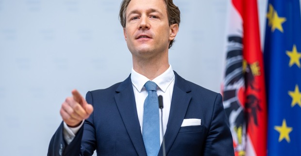 Gernot Blümel steht vor einem Mikrofon, spricht und deutet mit dem Zeigefinger in die Kamera. Hinter ihm österreichische und europäische Flagge.