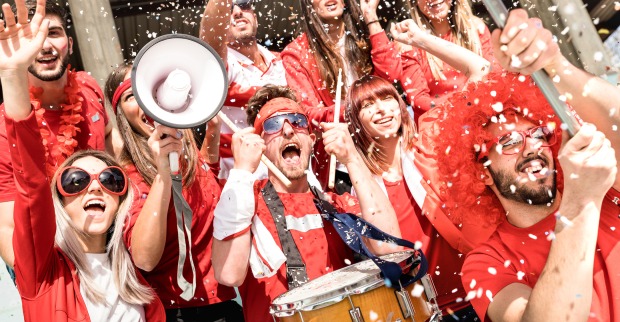 Eine Gruppe von Fußballfans feuert ihr Team mit Instrumenten und einem Megaphon in einem Konfetti-Regen stehend an