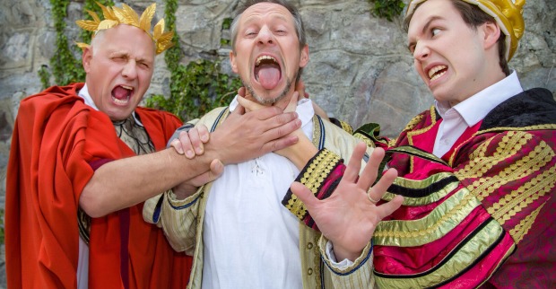 Ein Mann in einem Caeser-Kostüm und ein zweiter in der Verkleidung eines Königs würgen einen dritten im mittelalterlichen Outfit