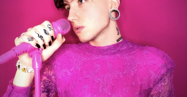 Ian Jules in einem pinken Kleid und mit einem Blumenkranz auf dem Kopf vor einem Mikrophon