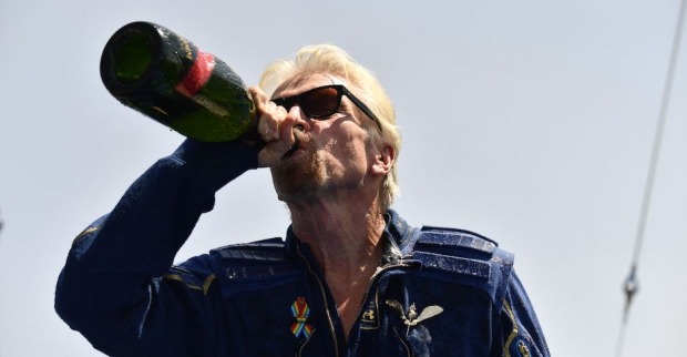 Richard Branson trägt eine Sonnenbrille und einen dunkelblauen Space-Overall. Er trinkt aus einer Flasche Champagner