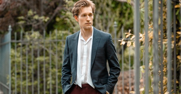 Der junge Musiker Simon Stadler steht, in einen modernen Anzug gekleidet, vor einem umzäunten Park