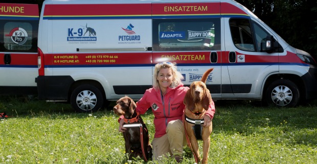 Alexandra Grunow kniet, flankiert von ihren zwei Hunden, auf einer Wiese vor dem Einsatzwagen des K9 Pro Vermisstensuchevereins