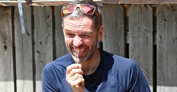 Herbert Kickl macht einen Corona-Selbstest. Er fährt mit einem Wattestäbchen in die Nase und zieht eine Grimasse. Kickl trägt eine Sonnenbrille am Kopf und sitzt vor einer Holzwand. Das Foto ist bei einer Wanderung entstanden.