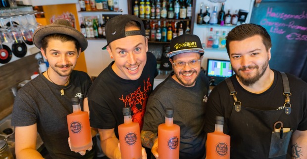 Vier Barkeeper stehen hinter einem Tresen und halten Flaschen mit einem orangen Inhalt in die Kamera