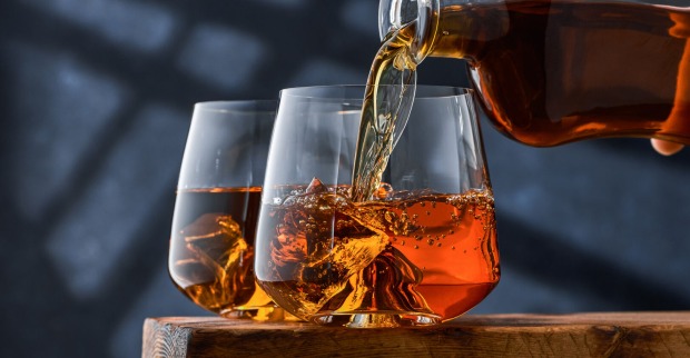 Zwei elegante Gläser Whiskey stehen auf einem Tisch, das sich im Vordergrund befindende wird aus einer Flasche befüllt