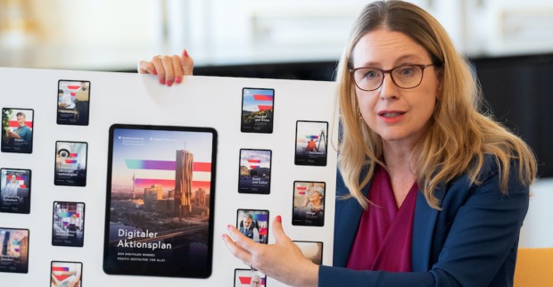 Margarete Schramböck hält einen großformatigen Ausdruck. Darauf zu sehen sind Fotos von Handy- und Tablet-Displays mit dem Slogan "Digitaler Aktionsplan".
