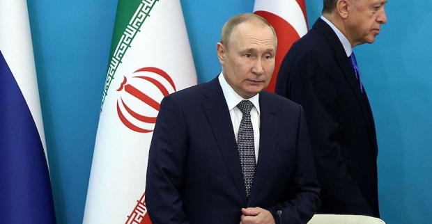 Wladimir Putin vor der russischen, iranischen und türkischen Fahne. Er wirkt überrascht und angeschlagen. Hinter seinem Rücken geht der türkische Präsident Erdogan vorbei.