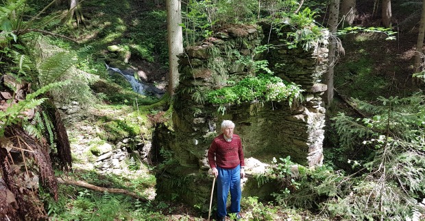 Ein alter Mann mit Gehstock steht vor der Ruine eines Partisanenunterschlupfs in einem Wald