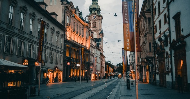 Blick in die Grazer Herrengasse bei Dämmerung. Einzelne Geschäfte sind beleuchtet, die Straßenbeleuchtung ist dunkel