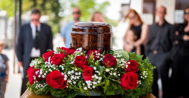 Nahaufnahme einer Urne mit Blumenkranz vor einer sich im Hintergrund befindenden Trauergemeinschaft.