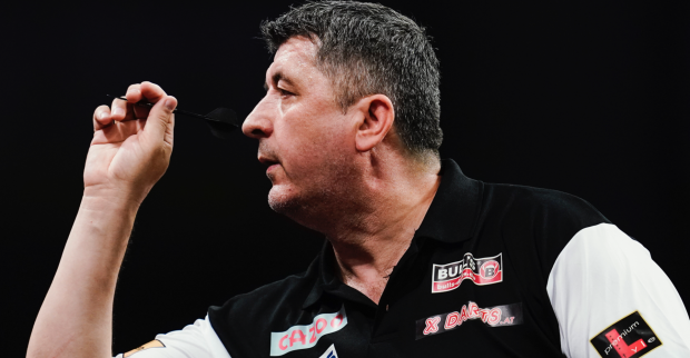 Nahaufnahme des Darts-Profis Mensur Suljović, der ein schwarzes Hemd mit weißen Ärmeln trägt und im Begriff ist, einen Dartspfeil zu werfen
